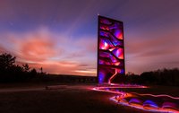 Der beleuchtete Architektur-Turm "Rostiger Nagel" bei Dämmerung mit Lichtspuren am Boden und bewaldetem Hintergrund